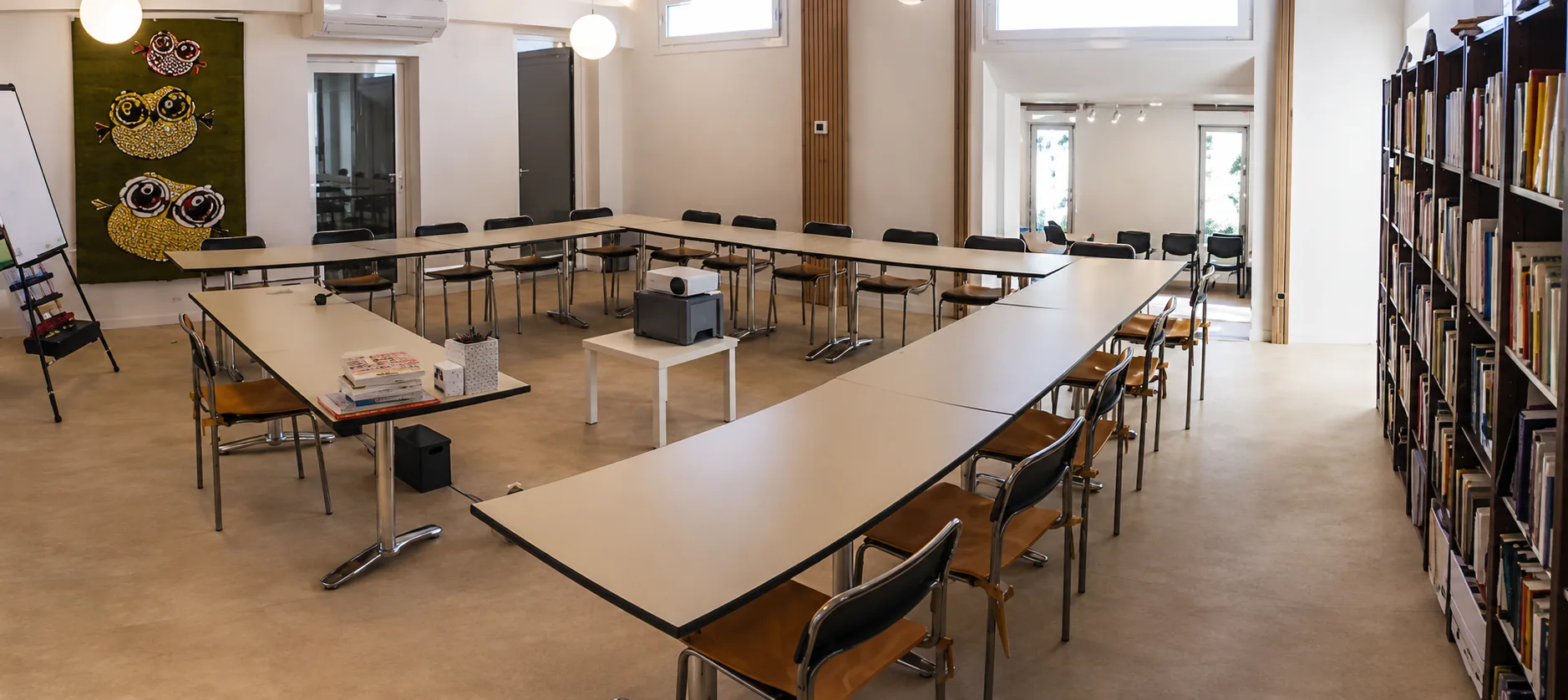 Een licht, modern trainingslokaal met U-vormig opgestelde tafels, een beamer en boekenkasten langs de wand.
