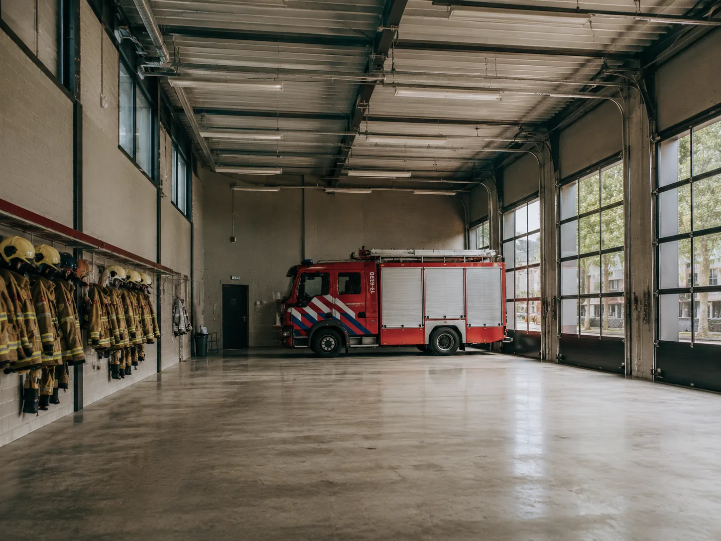 Het interieur van een Nederlandse brandweerkazerne &mdash; een rode brandweerwagen geparkeerd in een schone garage, brandweerjassen aan de linkerwand, daglicht door grote ramen.