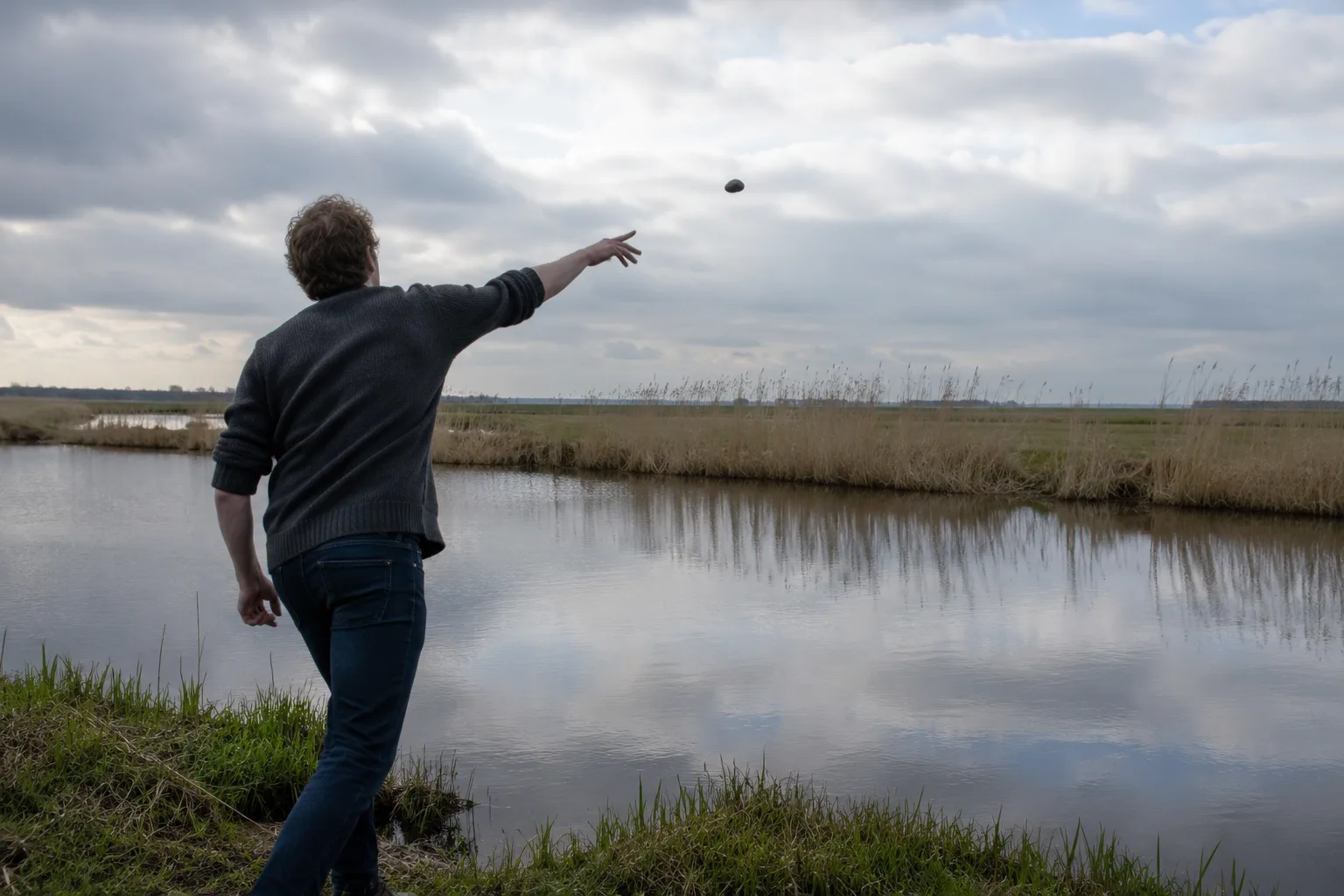 Een persoon van achteren gezien staat aan de rand van een polder-sloot en gooit in een bovenhandse worp een steen het water in; de steen hangt in de lucht boven het stille wateroppervlak.